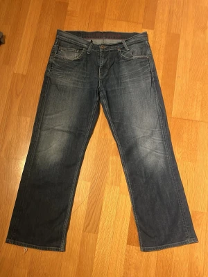 Blå bootcut  jeans med slitningar - Säljer ett par blå jeans med raka ben och diskreta slitningar på framsidan. Jeansen har klassiska fem fickor och knappstängning. Materialet är denim och färgen är mörkblå med ljusare partier. Perfekt för dig som gillar en avslappnad stil.