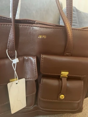 Brun axelväska från JW PEI - Snygg brun axelväska från JW PEI i veganskt läder med två stora fickor framtill och guldfärgade detaljer. Väskan har dubbla handtag och justerbar axelrem. Perfekt för dig som vill ha plats för allt och gillar en stilren men trendig look.