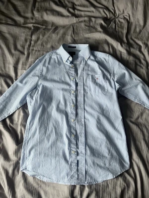 Blåvit randig skjorta från Gant - Klassisk blåvit randig skjorta från Gant med knappar framtill och liten logga på bröstfickan. Skjortan har lång ärm och är tillverkad i mjuk bomull. Perfekt för en clean och stilren look.