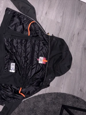 Svart pufferjacka med huva från Parajumpers - Snygg svart pufferjacka från Parajumpers med quiltad front och mjuka ärmar. Jackan har huva, dragkedja framtill och orangea detaljer på insidan. Perfekt för kalla dagar och riktigt skön att bära. Skulle villa säga bästa vår jackan! DE STÅR ATT DE ÄR M MEN DE ÄR XS! Får med kvitto på köpet! Ny pris 3900 kr ungiför.