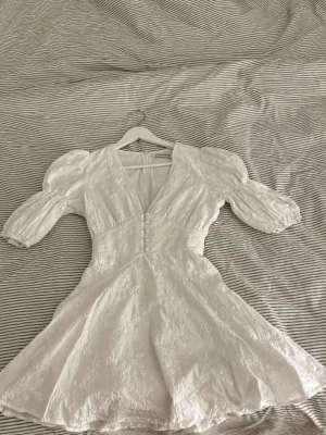 Vit kort klänning från Adoore - Superfin vit klänning från Adoore med v-ringning, puffade korta ärmar och dekorativa knappar framtill. Klänningen har broderade detaljer och en dragkedja i ryggen. Perfekt för studenten eller om du vill vara lite extra fin! Endast testad, säljer då den tyvärr är för liten för mig 