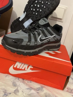 Nike grå och svarta sneakers - Snygga Nike sneakers i grått och svart med vit swoosh. Skorna har en sportig design med mesh och syntetmaterial, samt robust sula för bra grepp. Perfekta för dig som gillar streetwear och vill ha en modern look.