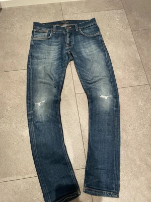 Blå jeans från Nudie Jeans - Säljer ett par blå jeans från Nudie Jeans med slitningar på knäna och klassisk femficksdesign. Jeansen har orangea sömmar och snyggt tvättad look med ljusare partier. Materialet är denim och passformen är rak. W32 L34 