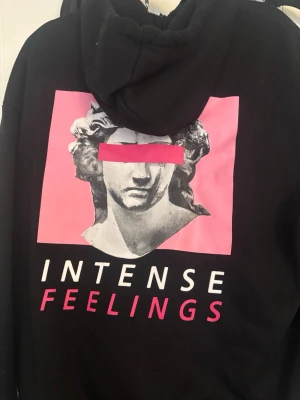Svart oversize hoodie med rosa tryck - Snygg svart oversize hoodie med huva och ett stort grafiskt tryck på bröstet i rosa och vitt. Texten 'INTENSE FEELINGS' står under en staty-inspirerad illustration. Hoodien har en avslappnad passform och är gjord i mjukt bomullsmaterial.