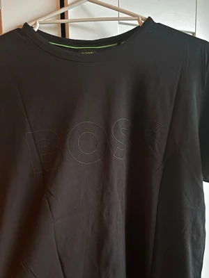 Svart t-shirt från BOSS - Snygg svart t-shirt från BOSS med diskret ton-i-ton BOSS-logga på bröstet. Klassisk rund halsringning och korta ärmar. Perfekt basplagg med stilren design, tillverkad i mjukt bomullsmaterial.