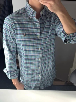Rutig skjorta från Ralph Lauren - Klassisk rutig skjorta från Ralph Lauren i blå, grön och vit färgskala. Skjortan har lång ärm, knappar framtill och button-down krage. Tillverkad i mjuk bomull och har den ikoniska broderade loggan på bröstet. Perfekt för en avslappnad men stilren look.
