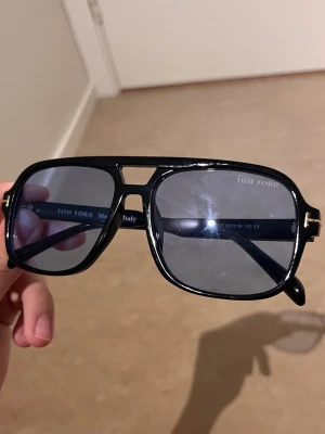 Svarta Tom Ford pilotbågar solglasögon - Snygga svarta solglasögon från Tom Ford med klassisk pilotform och mörka glas. Ramen är i blank svart plast med diskreta gulddetaljer vid gångjärnen. Perfekta för dig som vill ha en stilren och trendig look.