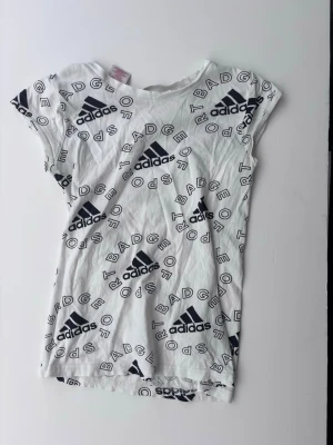 Vit Adidas t-shirt med loggor - Säljer en vit t-shirt från Adidas med svart logotyp och textmönster över hela plagget. Tillverkad i mjukt och lätt material, perfekt för träning. Storlek 164/XS 