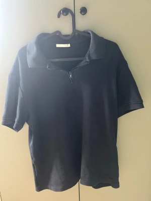 Svart polotopp med half zip från Zara - Svart polotopp från Zara i mjukt bomullsmaterial. Toppen har korta ärmar, klassisk krage och en half zip-dragkedja framtill som ger en sportig touch. Enkel och clean design som är lätt att matcha.