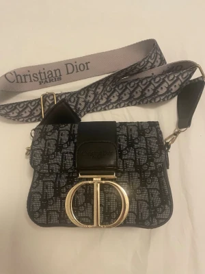 svart handväska - Snygg svart handväska med ikoniskt monogrammönster och bred axelrem med logotyp. Väskan har ett stort guldfärgat  framtill och detaljer i läder. Perfekt accessoar för dig som gillar lyxiga designer och statement-detaljer.