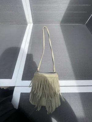 Beige axelväska med fransar - Snygg ljus grön axelväska i mockaimitation med långa fransar på framsidan. Väskan har en smal axelrem och stängs med dragkedja. Perfekt för dig som gillar boho-stil och vill ha en unik accessoar till din outfit.