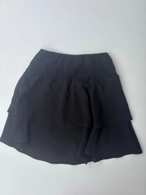 Svart volangkjol från ONLY - Supersnygg svart kortkjol från ONLY med bred smock i midjan och dubbla volanger. Kjolen har en flowy passform och är perfekt för dig som gillar en enkel men trendig stil. Tillverkad i mjukt tyg som känns skönt mot huden.