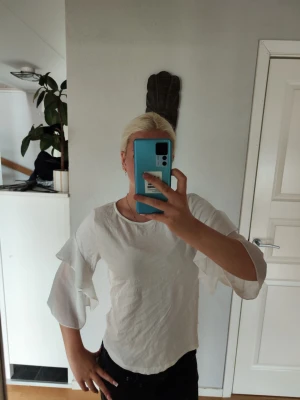 Vit blus med volangärmar från Lindex - Snygg vit blus från Lindex med trekvartsärm och volangdetaljer längs ärmarna. Blusen har rund halsringning och är gjord i ett lätt, luftigt material som ger en skön känsla. Perfekt att styla med jeans eller kjol för en trendig look.