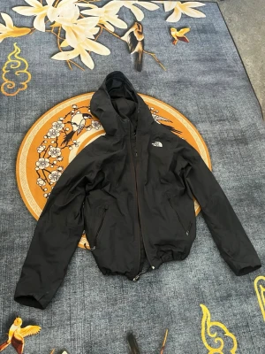 Svart vindjacka från The North Face - Snygg svart vindjacka från The North Face med huva och dragkedja framtill. Jackan har två fickor med dragkedja och elastisk nederkant. Perfekt för blåsiga dagar och enkel att matcha med olika outfits. Logga på bröstet och ryggen ger en sportig vibe. Storlek M. Pris kan diskuteras.
