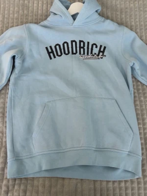 Ljusblå hoodie från Hoodrich - Snygg ljusblå hoodie från Hoodrich med stor magficka och svart logga tryckt över bröstet. Klassisk huva och ribbade muddar vid ärmslut och nederkant. Perfekt för en chill och avslappnad stil.