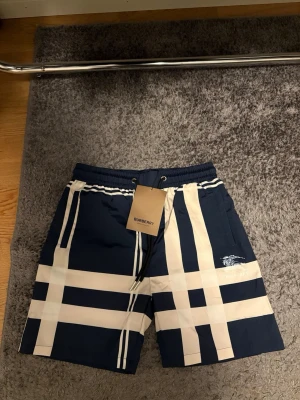 Marinblå rutiga shorts från Burberry - Oanvända shorts från Burberry i marinblått med stort rutmönster i beige och vitt. Perfekta badshorts nu till sommaren om man vill glida runt med stil på stranden men samtidigt kunna bära de vanligt till vardags också. M i storlek men passar S också!