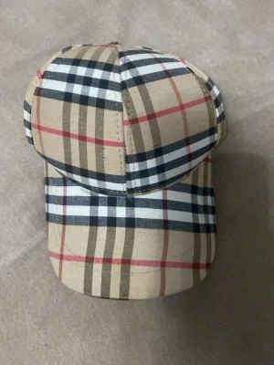 Burberry  - Säljer en beige keps med klassiskt rutigt mönster i svart, vitt och rött. Kepsen har böjd skärm och justerbart metallspänne bak för perfekt passform. Tillverkad i bomullstyg och har en riktigt clean och tidlös look.