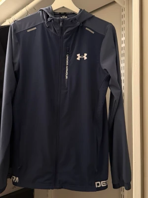 Blå vindjacka från Under Armour - Snygg marinblå vindjacka från Under Armour med huva och dragkedja framtill. Jackan har vita logodetaljer, perforerade partier på sidorna för ventilation och en bröstficka med dragkedja. Tillverkad i lätt och funktionellt material, perfekt för aktiva dagar.