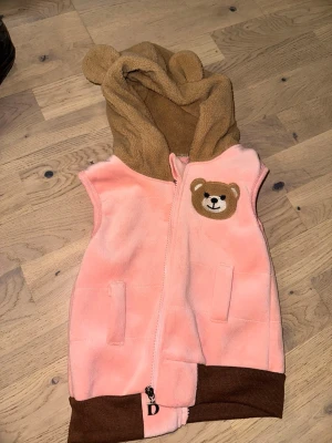 Rosa fleeceväst med björndetalj - Supersöt rosa fleeceväst med bruna muddar och huva med björnöron. På bröstet finns en broderad björn och västen har dragkedja framtill samt två fickor. Perfekt för dig som vill ha en mysig och lekfull stil.Rosa fleeceväst med björndetalj