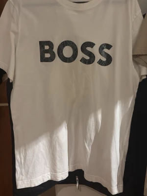 Vit BOSS t-shirt med tryck - Vit t-shirt från BOSS med stort svart BOSS-tryck på bröstet. Klassisk rund halsringning och korta ärmar. Perfekt för en clean och stilren look.