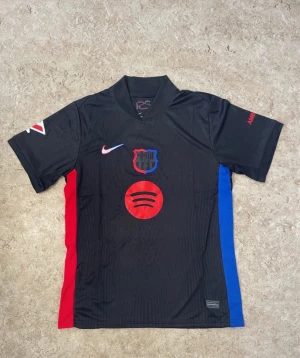 Svart FC Barcelona matchtröja Nike - Säljer en svart FC Barcelona matchtröja från Nike med klubbmärke och Spotify-logga på bröstet. Tröjan har röda och blå sidopaneler, korta ärmar och tryck på både fram- och baksida. Perfekt för dig som älskar fotboll och vill ha en snygg supportertröja.