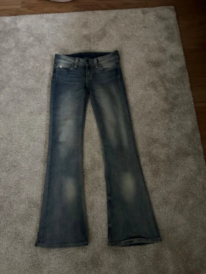 Blå bootcut jeans från Cheap Monday - Säljer ett par blå bootcut jeans från Cheap Monday med slitna detaljer och klassisk femficksmodell. Jeansen har en ljus tvätt och utsvängda ben, perfekta för en avslappnad och trendig look. Storlek 26/30