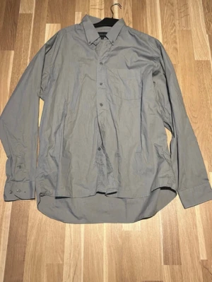 Grå långärmad skjorta - En stilren grå skjorta med klassisk krage och knäppning framtill. Skjortan har lång ärm med manschett och en bröstficka. Perfekt för en clean och enkel look.