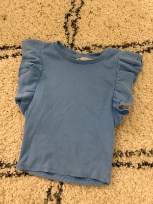 Blå ribbad topp med volang från Zara - Supersöt ljusblå topp från Zara med ribbad struktur och volangdetaljer på ärmarna. Perfekt till jeans eller kjol för en fräsch look. Materialet känns mjukt och stretchigt, och modellen är lite croppad. (Skriv för fler bilder💕)