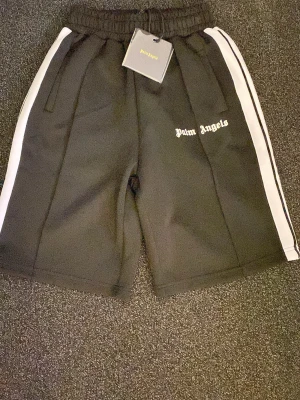 Svarta shorts från Palm Angels - Palm Angels svarta shorts med vita sidoränder och logga broderad på vänster ben. Elastisk midja med snörning och sportig look. Perfekta för dig som gillar streetwear och vill ha en clean men ändå statement stil.