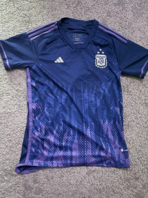 Lila Argentina matchtröja Adidas - Snygg lila matchtröja från Adidas med Argentinas landslagsmärke och två stjärnor på bröstet. Tröjan har korta ärmar, rund hals och ett mönster i olika lila nyanser. Tillverkad i lätt och ventilerande funktionsmaterial, perfekt för träning eller match. Säljer då den var för liten och alltså näst intill aldrig används och är som ny.