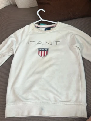 Vit hoodie från GANT med logga - Vit hoodie från GANT med klassisk logotyp och emblem på bröstet. Tröjan har långa ärmar och rund halsringning. Materialet är mjukt och bekvämt, perfekt för en avslappnad stil. Passar dig som gillar stilrena och tidlösa plagg.