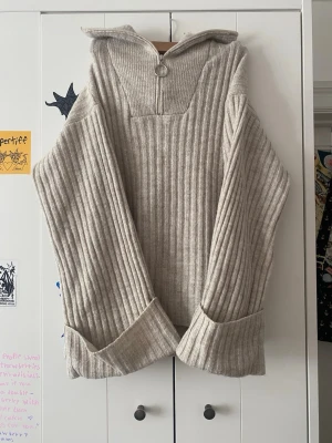 Beige ribbstickad tröja med dragkedja - Säljer en beige ribbstickad tröja med hög krage och halv dragkedja med ringdetalj. Tröjan har långa, vida ärmar med breda uppvikta muddar och en avslappnad passform. Perfekt för lager på lager och riktigt mysig till hösten.