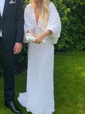Vit långklänning med paljetter - Elegant vit långklänning med djup v-ringning och vida ärmar. Klänningen är täckt av glittriga paljetter. Perfekt för speciella tillfällen.