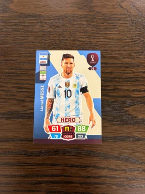 Lionel Messi fotbollskort VM 2022 - Samlarfotbollskort från FIFA World Cup Qatar 2022 med HERO-design. Kortet har färgerna blå, vit och röd med detaljer i guld och svart. 