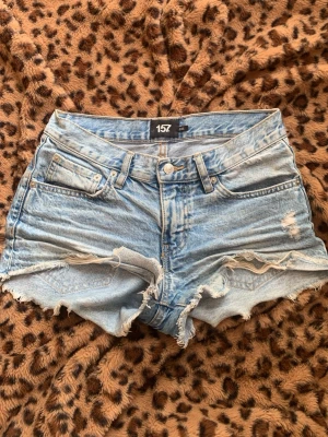 Ljusblå denimshorts från 157 - Säljer ett par ljusblå denimshorts från 157 med hög midja och råa, fransiga kanter. Shortsen har klassisk femficksdesign, slitna detaljer och knappgylf. Perfekta för sommaren och ger en avslappnad vibe.