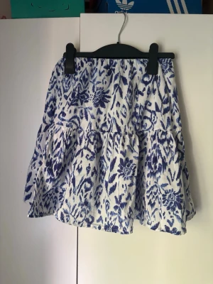 Blåvit blommig kjol från Gina Tricot - Supersöt kort kjol från Gina Tricot i blått och vitt med blommigt mönster. Kjolen har volang och resår i midjan för en bekväm passform. Perfekt för dig som gillar en fräsch och somrig stil.