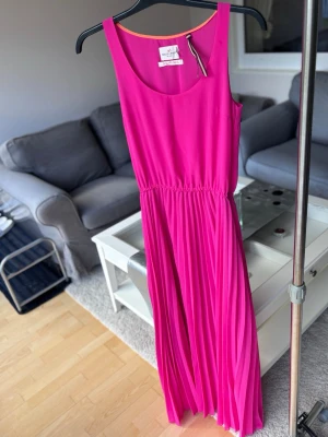 Plisserad långklänning i stark rosa - Snygg långklänning i stark rosa med plisserad kjol och markerad midja. Klänningen är ärmlös och har en enkel, elegant design som ger ett modernt intryck. Perfekt för dig som vill sticka ut med färg och stil.
