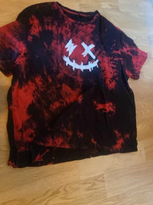 Röd och svart tie-dye t-shirt - Cool t-shirt med röd och svart tie-dye effekt och ett vitt smileytryck med korsade ögon på bröstet. Klassisk passform och korta ärmar, perfekt för dig som gillar streetwear och vill sticka ut lite extra.