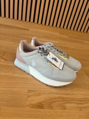 Grå sneakers från U.S. Polo Assn. - Snygga grå sneakers från U.S. Polo Assn. med detaljer i guld och ljusrosa. Skorna har klassisk snörning och en vit sula. Märkets logga syns på sidan och hälen har en rosa kontrast. Perfekta för en trendig vardagslook. Helt nya aldrig använt dom.
