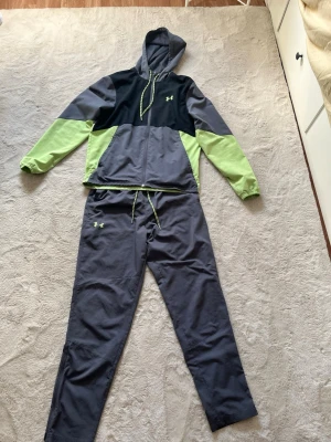 Under Armour träningsset grå/svart/grön - Säljer ett snyggt träningsset från Under Armour med hoodie och byxor. Setet är i grått, svart och ljusgrönt med dragsko i midjan och på huvan. Perfekt för gymmet eller löprundan. Under Armour-logga på både tröja och byxa.