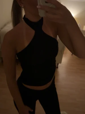 Svart oneshoulder topp med hög hals - Snygg svart topp med oneshoulder-design och hög hals. Toppen är ärmlös och sitter tight mot kroppen, perfekt för en trendig look. Materialet känns mjukt och stretchigt, vilket gör den bekväm att bära. Passar dig som gillar stilrena och moderna plagg.