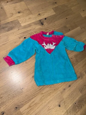 Blå och cerise pyjamas med fågelmotiv - Mysig pyjamas i mjuk frotté med långa ärmar. Färgen är klarblå med cerise detaljer vid hals och ärmslut samt ett broderat fågelmotiv framtill. Perfekt för sköna hemmakvällar.Blå och cerise pyjamas med fågelmotiv