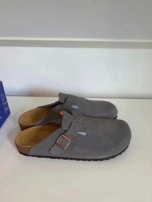 Gråa Birkenstock tofflor i mocka - Säljer ett par gråa Birkenstock tofflor i mjuk mocka med justerbart spänne i kopparfärg. Tofflorna har en klassisk slip-in design och bekväm innersula i kork. Perfekta för chill dagar hemma eller när du vill ha något skönt på fötterna.