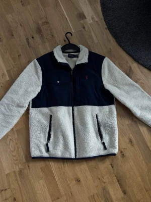 Teddyjacka Polo Ralph Lauren - Teddyjacka från Polo Ralph Lauren i marinblått och vitt. Jackan har mjukt, fluffigt teddy-material, dragkedja framtill, bröstficka med knapp och två sidofickor med dragkedja. Klassisk krage och broderad logga på bröstet. Perfekt för kyliga dagar.