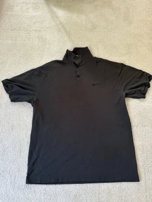 Svart polotopp från Nike - Snygg svart polotopp från Nike med korta ärmar och diskret logga på bröstet. Toppen har en hög krage med knappar framtill och är tillverkad i ett mjukt, lätt material som känns skönt mot huden. Perfekt för en clean och sportig look.