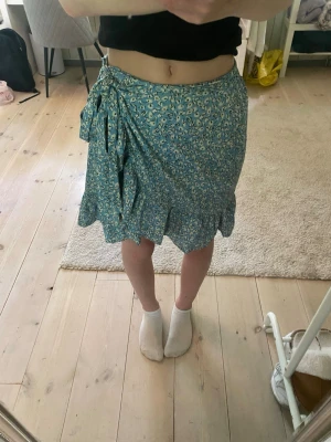 Blommig blå omlottkjol med volang - Supersöt blå omlottkjol med blommigt mönster och volangkant. Kjolen knyts i sidan med en stor rosett och har en lätt och luftig känsla, perfekt för varma dagar. Passformen är normal och kjolen slutar ovanför knät. Säljes då den ej kommer till användning