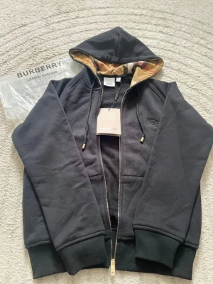 Svart hoodie från Burberry med dragkedja - Snygg svart hoodie från Burberry med dragkedja och klassiskt rutigt foder i huvan. Hoodien har ribbade muddar, fickor framtill och guldfärgade detaljer på snörena. Perfekt för dig som vill ha en exklusiv och stilren look. Materialet är mjukt och bekvämt bomullstyg. Påsen kommer med.