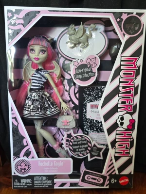 Monster High, Boo-riginal creeproduktion - Monster High Rochelle Goyle docka i originalförpackning. Dockan har rosa och blått hår, vingar och en svartvit klänning med mönster. Medföljer dagbok, dockställ och en liten väska. Förpackningen är svart, rosa och vit med Monster High-logga.