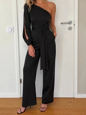 Svart jumpsuit one shoulder stl S - Svart jumpsuit med en axel och lång ärm med slits. Jumpsuiten har ett knytband i midjan. Polyester. Använd en gång, är i mkt bra skick. Inga defekter. Stl S.