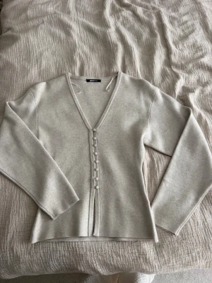 Ljusbeige kofta från Gina Tricot - Ljusbeige kofta från Gina Tricot med v-ringning och klädda knappar framtill. Koftan är tunnstickad och har långa ärmar. Perfekt att slänga över en topp för en clean och enkel look.